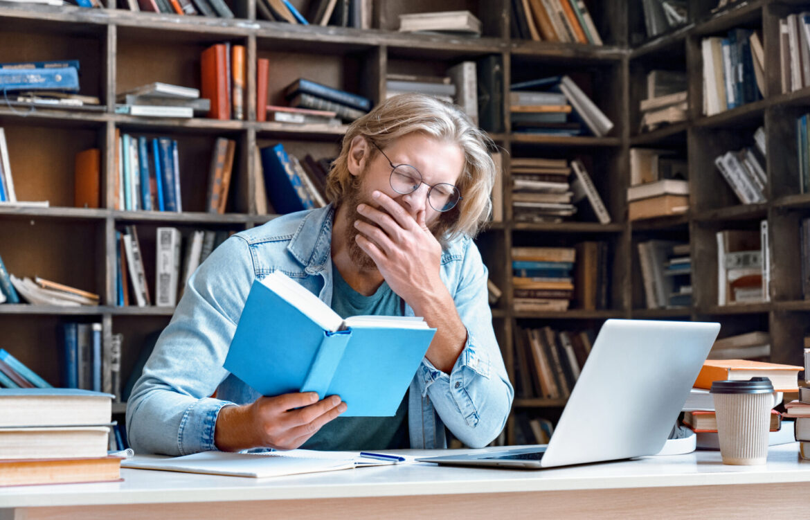 Bored tired sleepy guy university student yawning reading book studying in library feel lack of sleep of dull learning sit at desk preparing for college exam, fatigue of boring education concept