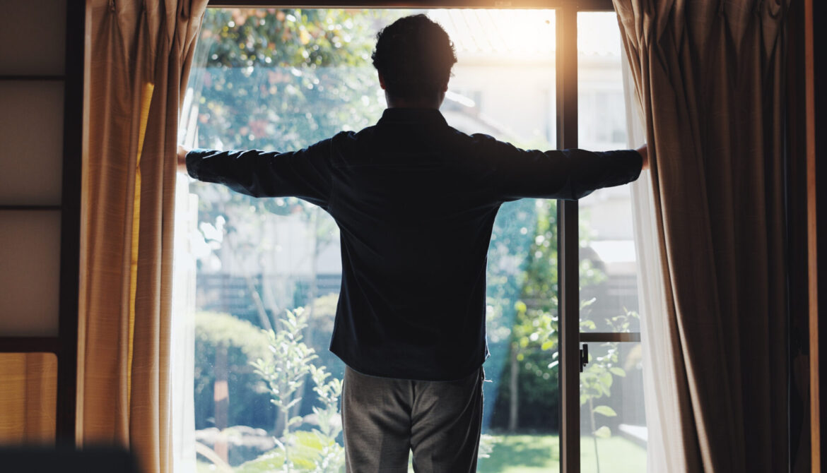 Morning, home and open curtain with man to start day with sunshine at vacation rental and accommodation. Bedroom, waking up and back with trip and holiday in Japan at window with light in hotel